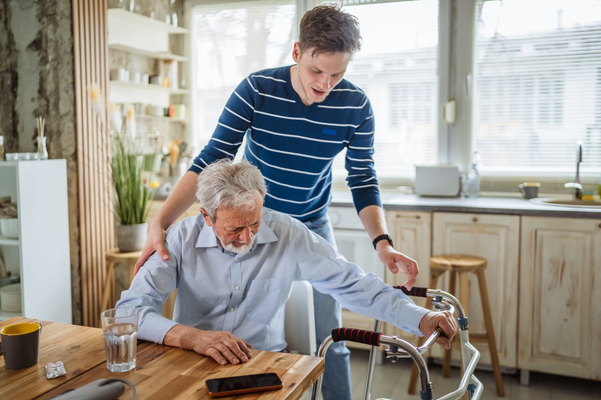 A young man supports a senior man using a mobility walker in a home kitchen